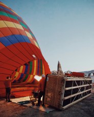 A colorful hot air balloon being inflated with flames at dawn. Two people stand beside the balloon's wicker basket on a barren landscape.