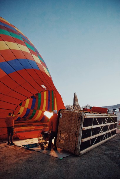 A colorful hot air balloon being inflated with flames at dawn. Two people stand beside the balloon's wicker basket on a barren landscape.