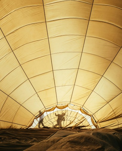 Inside view of a large, yellowed hot air balloon being inflated. Two silhouetted figures work, casting shadows on the fabric.