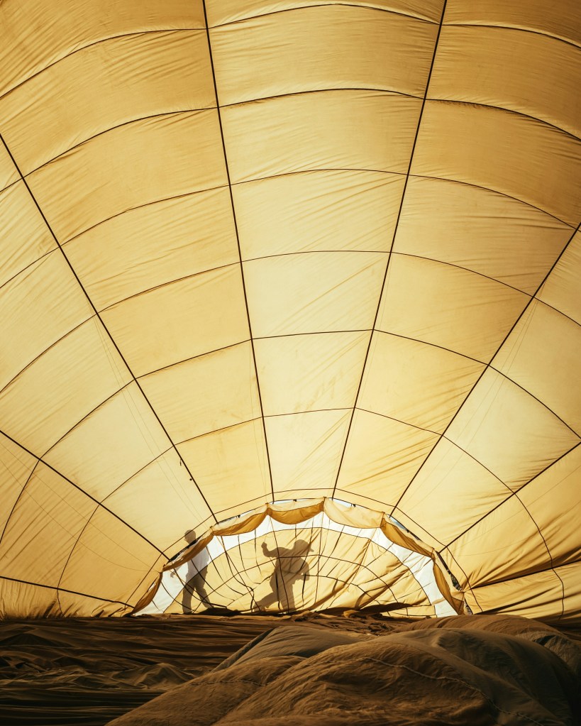 Inside view of a large, yellowed hot air balloon being inflated. Two silhouetted figures work, casting shadows on the fabric.