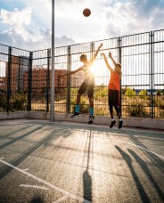 Two people jump to shoot a basketball on an outdoor court at sunset. Long shadows are cast on the ground, and a fence borders the court.
