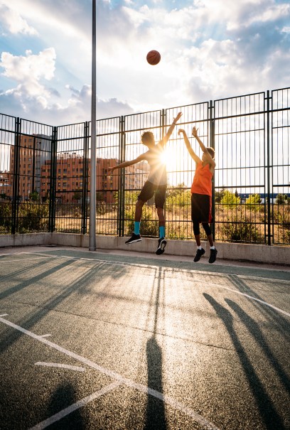 Two people jump to shoot a basketball on an outdoor court at sunset. Long shadows are cast on the ground, and a fence borders the court.