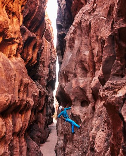 Climber in blue gear ascends a narrow, rugged red rock canyon under sunlight.