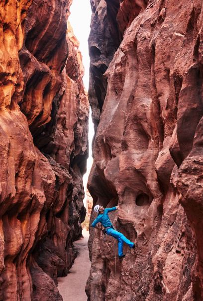 Climber in blue gear ascends a narrow, rugged red rock canyon under sunlight.