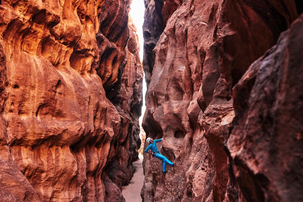Ein Kletterer in blauer Ausrüstung steigt im Sonnenlicht eine enge, zerklüftete Schlucht aus rotem Felsen hinauf.