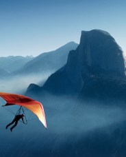 A person hang glides with a bright orange and red wing above misty mountains, including the iconic Half Dome, under a clear blue sky.
