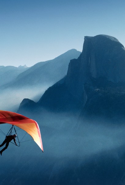 A person hang glides with a bright orange and red wing above misty mountains, including the iconic Half Dome, under a clear blue sky.