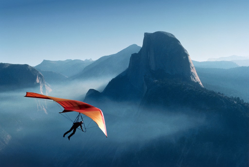 Eine Person gleitet mit einem leuchtend orangefarbenen und roten Flügel über den nebligen Bergen, darunter der ikonische Half Dome, unter einem klaren blauen Himmel.