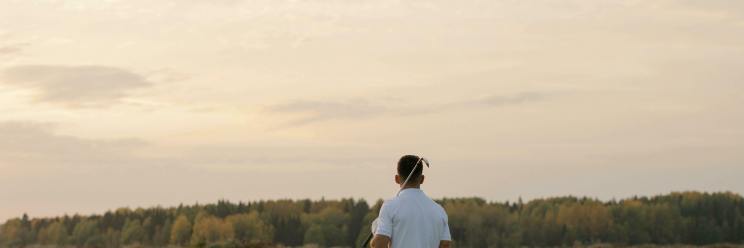 A person walks on a lush golf course carrying a club, wearing a white shirt and patterned pants. The sky is cloudy with a warm, serene sunset.