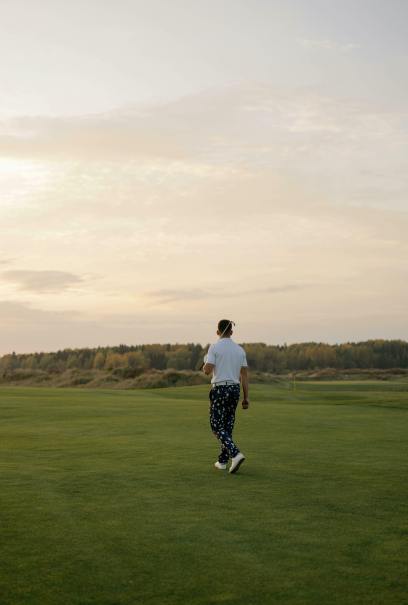 Eine Person geht mit einem Schläger, einem weißen Hemd und einer gemusterten Hose über einen üppigen Golfplatz. Der Himmel ist bewölkt mit einem warmen, heiteren Sonnenuntergang.