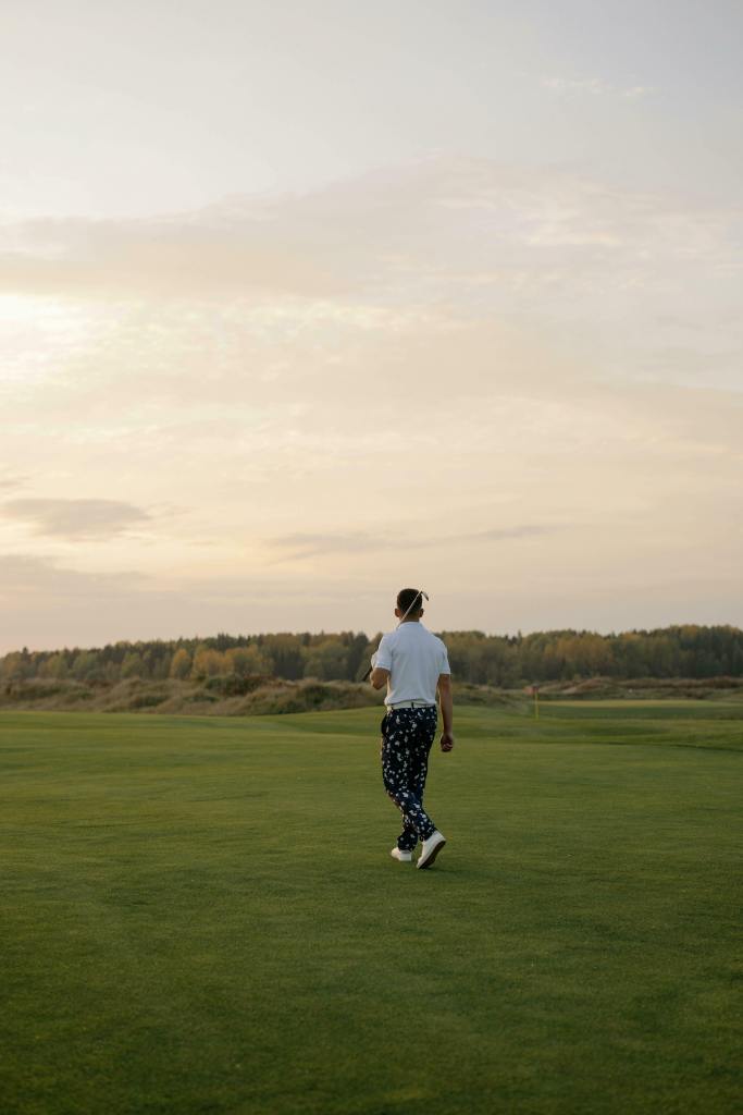 A person walks on a lush golf course carrying a club, wearing a white shirt and patterned pants. The sky is cloudy with a warm, serene sunset.