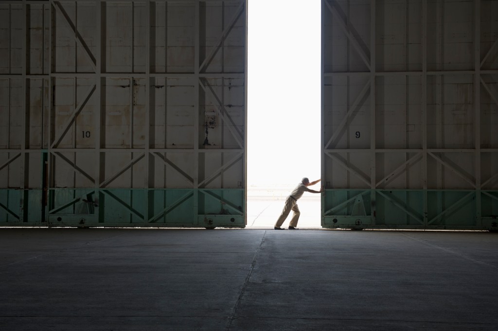A person pushes open a massive hangar door, revealing bright daylight outside.