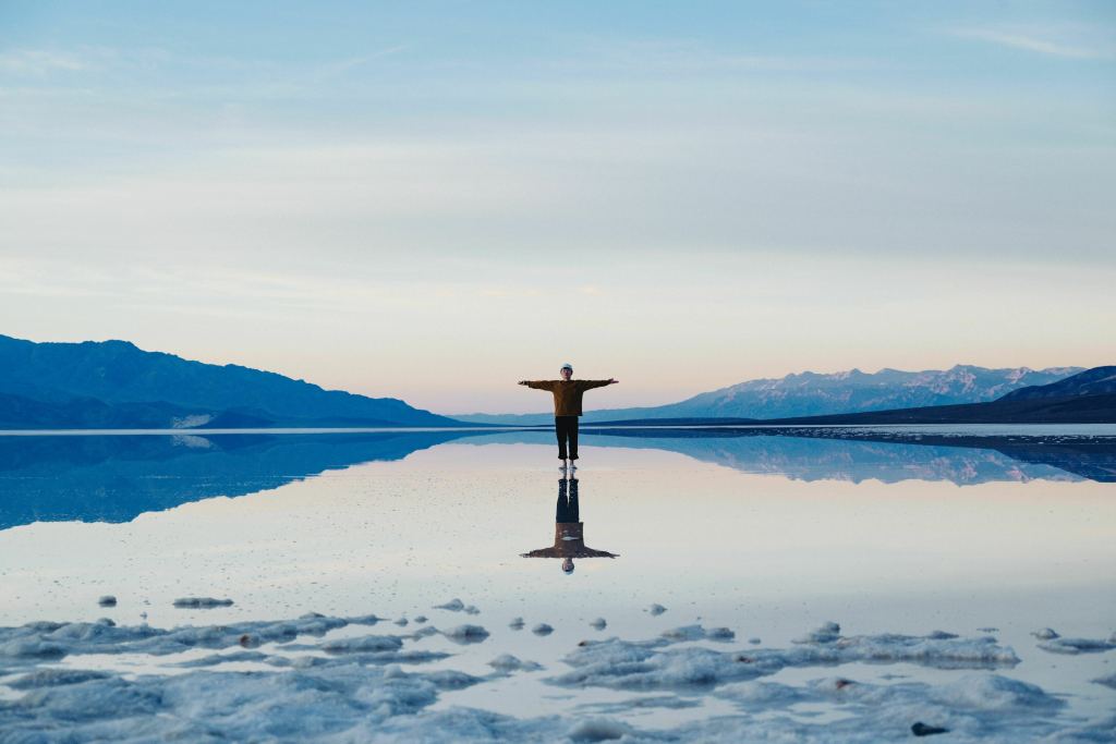 Une personne se tient debout, les bras tendus, se reflétant dans une étendue d'eau calme. Montagnes environnantes et ciel pastel.