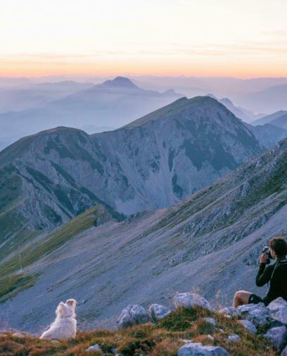 A person with a camera and a white dog sit on a rocky mountainside, overlooking vast mountain peaks at sunrise.