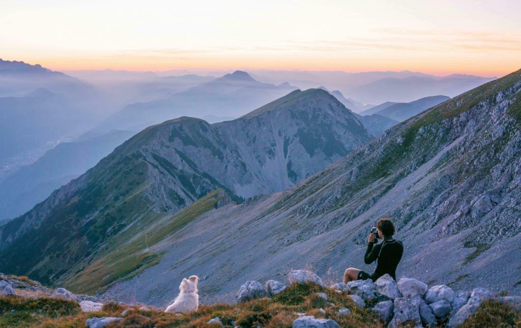 A person with a camera and a white dog sit on a rocky mountainside, overlooking vast mountain peaks at sunrise.