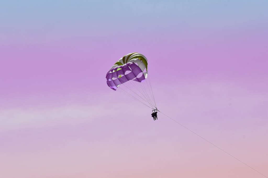A person parasails high in the sky under a purple and green parachute, set against a serene gradient of pink and blue hues.