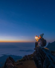 A person in warm clothing and a beanie sits on a rocky cliff at dawn, holding a glowing lantern. The sky is filled with stars, and the horizon is tinged with orange and blue.
