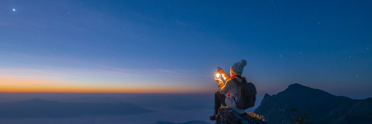 A person in warm clothing and a beanie sits on a rocky cliff at dawn, holding a glowing lantern. The sky is filled with stars, and the horizon is tinged with orange and blue.