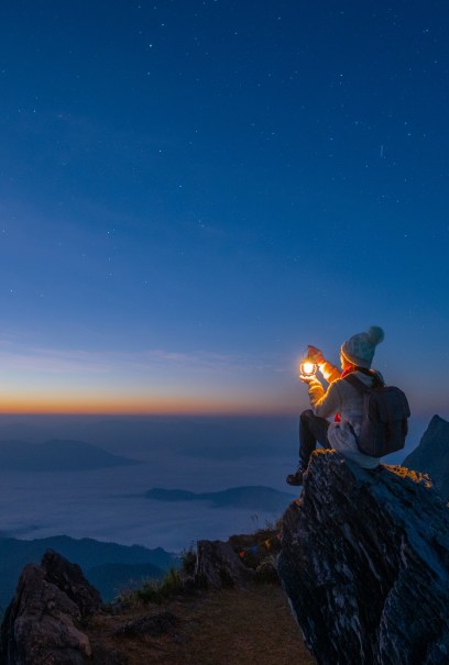 A person in warm clothing and a beanie sits on a rocky cliff at dawn, holding a glowing lantern. The sky is filled with stars, and the horizon is tinged with orange and blue.