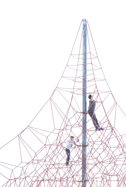 Two children climb a tall red rope structure against a clear sky.