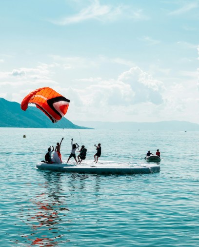 People enjoy parasailing from a floating platform on a serene lake. A vibrant red parachute is in the air, with mountains under a partly cloudy sky.