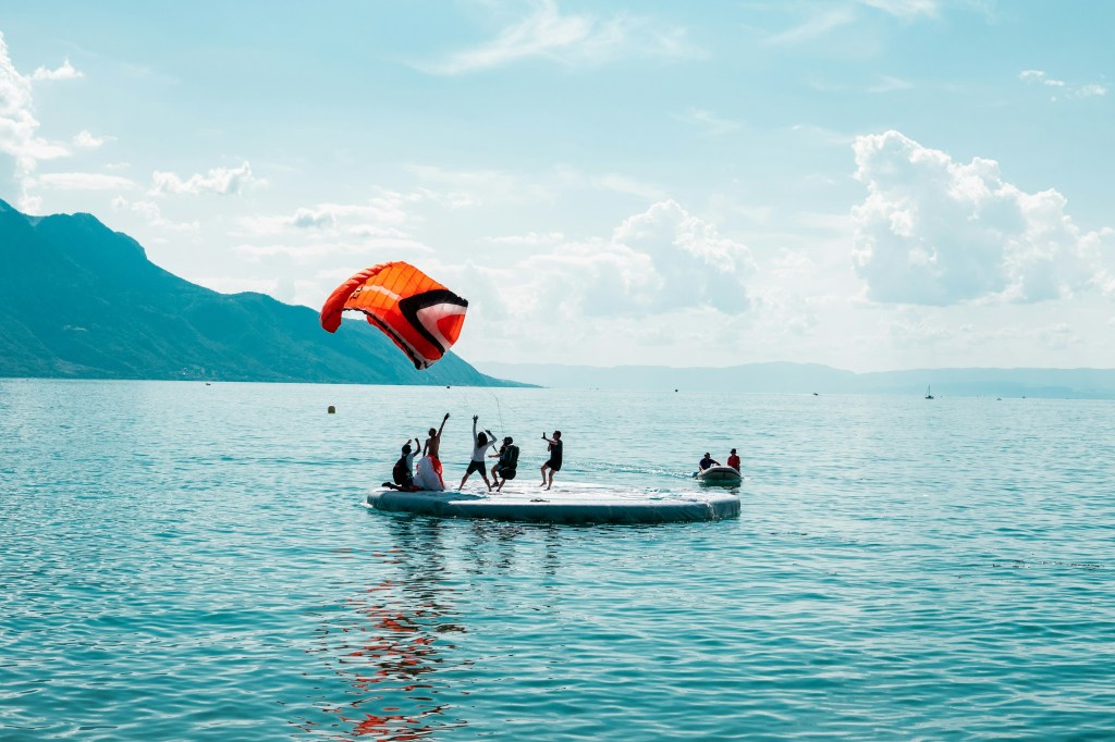 People enjoy parasailing from a floating platform on a serene lake. A vibrant red parachute is in the air, with mountains under a partly cloudy sky.