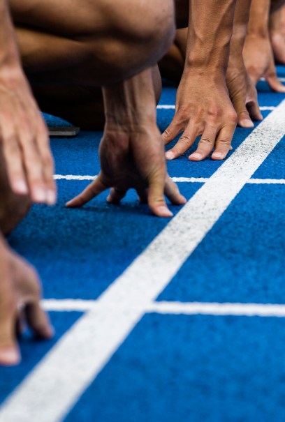 Nahaufnahme der Hände von Sprintern auf einer blauen Bahn, die an der Startlinie aufgereiht sind, bereit zum Rennen.