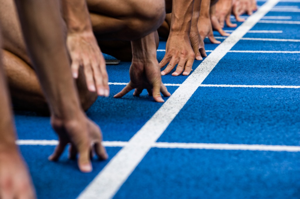 Gros plan sur les mains des sprinters sur une piste bleue, alignés sur la ligne de départ, prêts à courir.