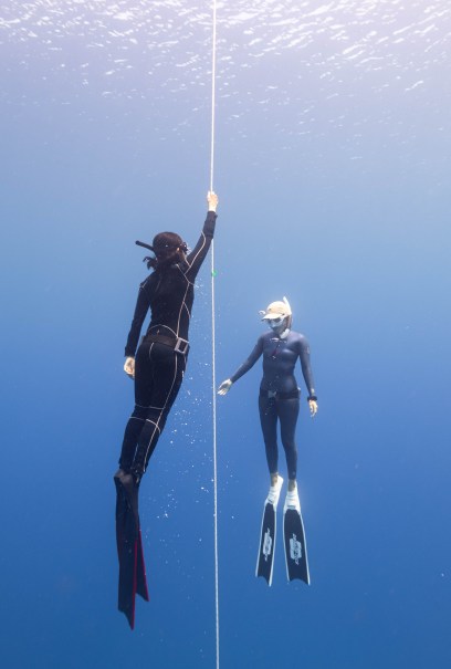 Two divers are underwater in clear blue water. One holds a guide rope, and both wear wetsuits, masks, and fins.