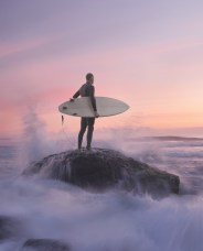 Surfer standing on a rock, holding a surfboard, gazing at waves crashing around during a serene sunset with pink and purple skies.