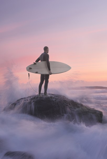 Surfer standing on a rock, holding a surfboard, gazing at waves crashing around during a serene sunset with pink and purple skies.