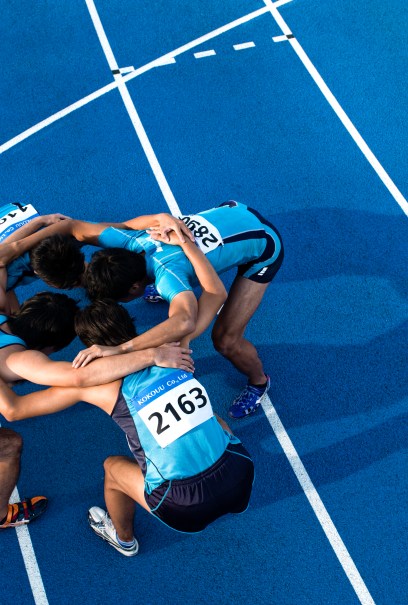 Four athletes in blue uniforms huddle closely on a bright blue running track.