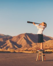A young girl stands on a stool, peering through a telescope towards distant mountains under a clear sky.