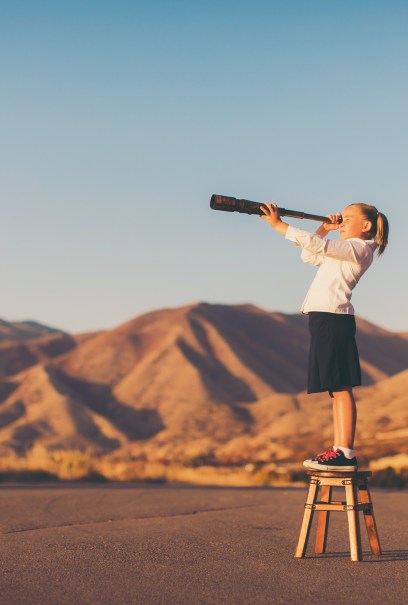 A young girl stands on a stool, peering through a telescope towards distant mountains under a clear sky.