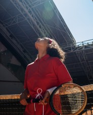 A tennis player in a red uniform stands by the net, head tilted back and eyes closed under a bright sky. She holds a racket.