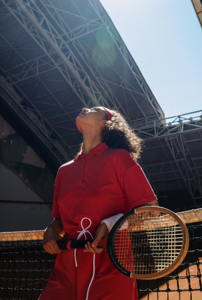 A tennis player in a red uniform stands by the net, head tilted back and eyes closed under a bright sky. She holds a racket.