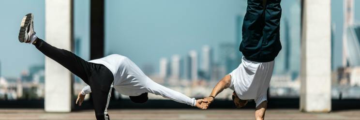 Zwei Breakdancer führen im Freien dynamische Stunts vor. Der eine steht im Handstand und hält die Hand des anderen, während sie sich nach vorne lehnen. Hintergrund ist die städtische Skyline.