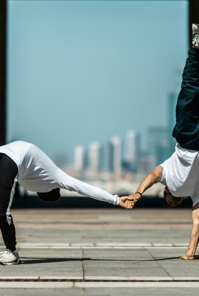 Two breakdancers perform dynamic stunts outdoors. One is in a handstand while holding the other's hand as they lean forward. Urban skyline background.