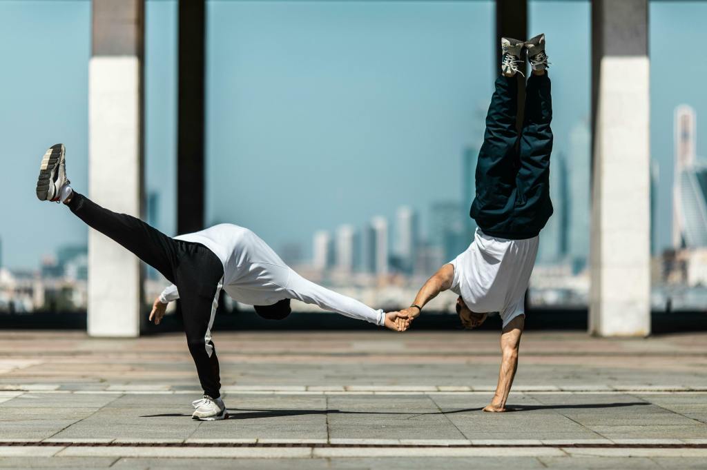 Two breakdancers perform dynamic stunts outdoors. One is in a handstand while holding the other's hand as they lean forward. Urban skyline background.