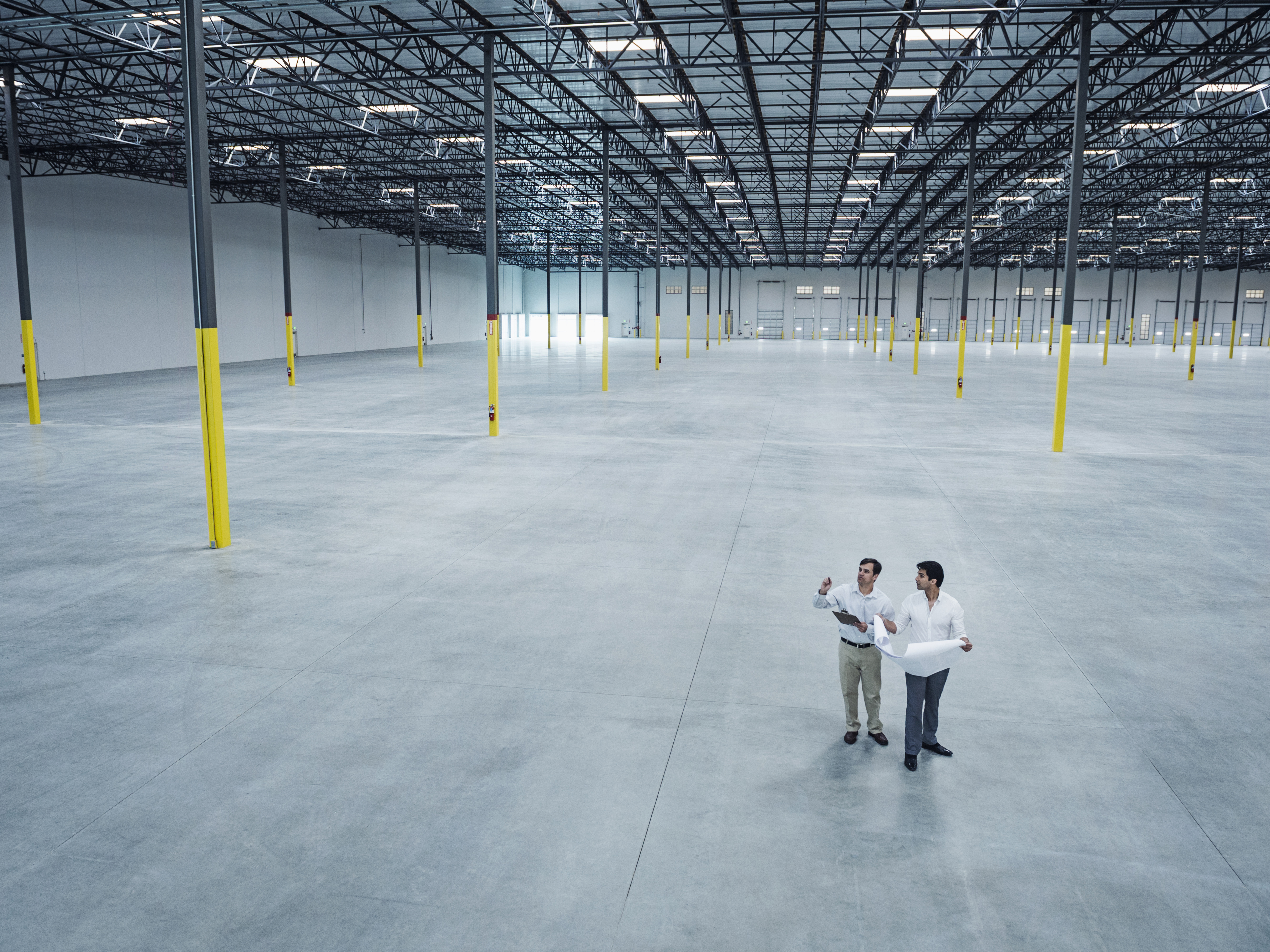 Two men in white shirts discuss plans in a spacious, empty warehouse with a high ceiling. Yellow pillars and large windows add depth to the scene.
