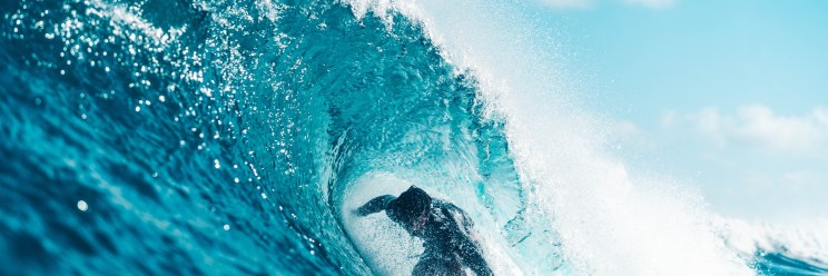 Surfer skillfully rides inside a powerful, curling blue wave under a bright sky.