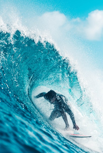 Surfer skillfully rides inside a powerful, curling blue wave under a bright sky.
