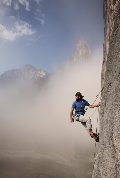 A man in climbing gear scales a steep rock face amidst misty mountains. The sky is partially cloudy.