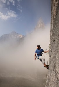 A man in climbing gear scales a steep rock face amidst misty mountains. The sky is partially cloudy.