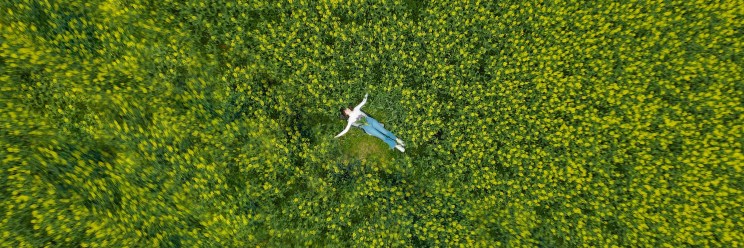 Aerial view of a person in a white shirt and blue jeans lying in a lush green field with outstretched arms, surrounded by vibrant yellow flowers.