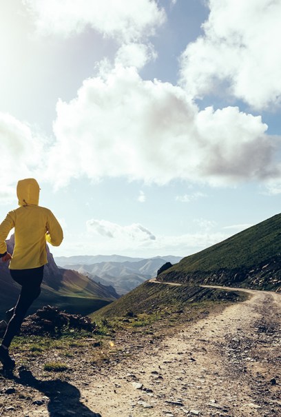 A person in a yellow jacket runs on a mountain trail, with rugged peaks and a clear, sunlit sky in the background.