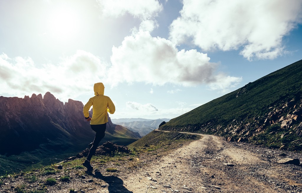 A person in a yellow jacket runs on a mountain trail, with rugged peaks and a clear, sunlit sky in the background.