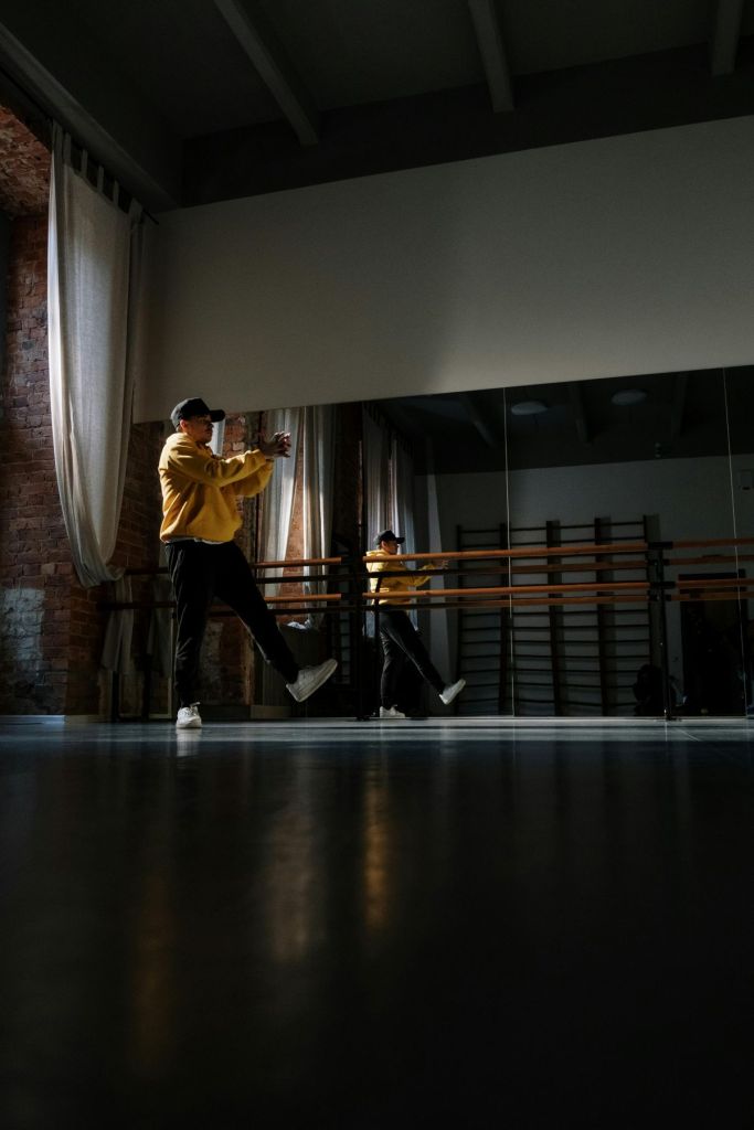 Person in a yellow hoodie, black cap, and dark pants practicing dance movements in a spacious studio with mirrored walls, exposed brick, and tall curtains. The dancer's reflection is visible in the mirror, and natural light creates reflections on the dark floor.